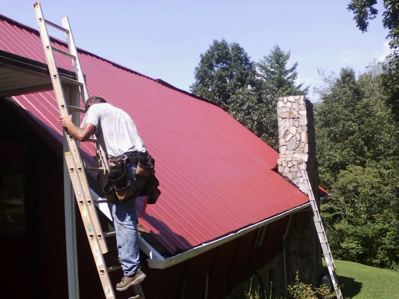 Roofer on a ladder beside a red metal roof and stone chimney — Soffit & Fascia Repair by Roofing Brothers in Veedersburg